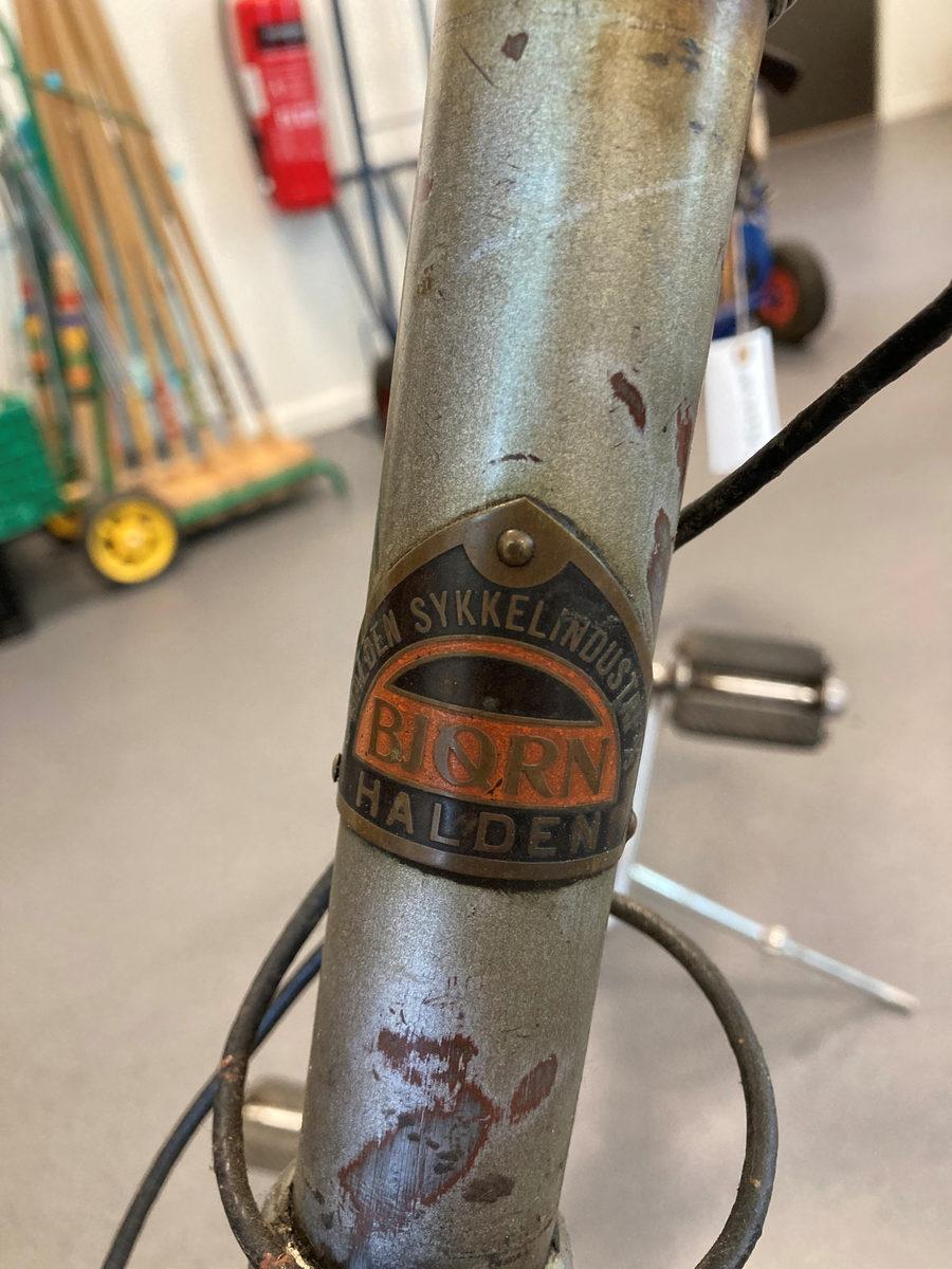 Herresykkel i metall, med sete av lær, lykt og bjelle. Påmontert verktøyeske i lær ved sete. Festet ved bagasjebrett 2 rødmalte halvsirkelformede kasser i tre, med en brannslange i hver.