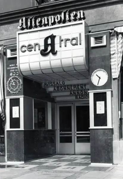 Viser et svarthvitt bilde av en blokk-aktig inngang, med et skilt som danner "terrasser" innover mot selve døren og lager en dybde-effekt til inngangen. Skiltet viser til Central A, hvor A-en står midstilt og i kontrast med Cen - Tral. "plattformene" viser: Avissalg, Abonnement, Annon-*kuttet av* og Radio.  