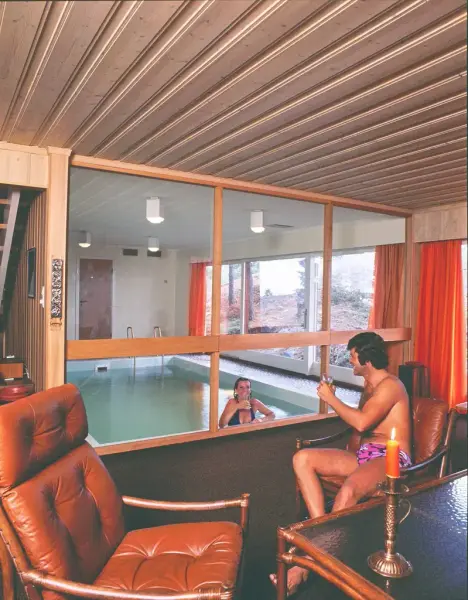 Bildet viser to rom separert med vinduer, det bakerste har ett innendørs svømmebasseng, og en glassvegg. Det andre rommet kan se ut som et kontor med to oransje skinnstoler, i den ene sitter en mann med typisk 70-talls hår som snakker med en dame på kanten av svømmebassenget på andre siden av vinduet.