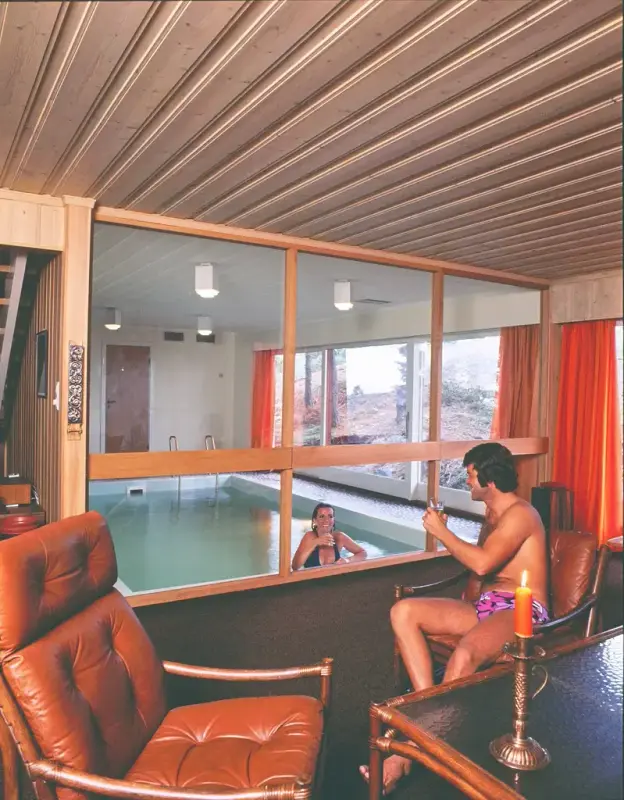 Bildet viser to rom separert med vinduer, det bakerste har ett innendørs svømmebasseng, og en glassvegg. Det andre rommet kan se ut som et kontor med to oransje skinnstoler, i den ene sitter en mann med typisk 70-talls hår som snakker med en dame på kanten av svømmebassenget på andre siden av vinduet.