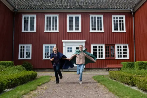 To barn med kapper løper i bastionshagen og man ser den gamle hovedinngangen på Herregården i bakgrunn. 