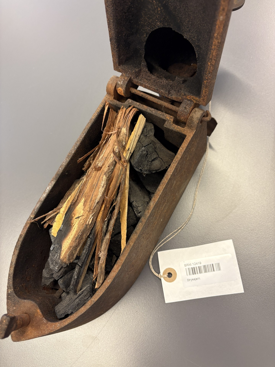 Båtformet strykejern av støpejern. Hengslet lokk med trehåndtak og pipe.