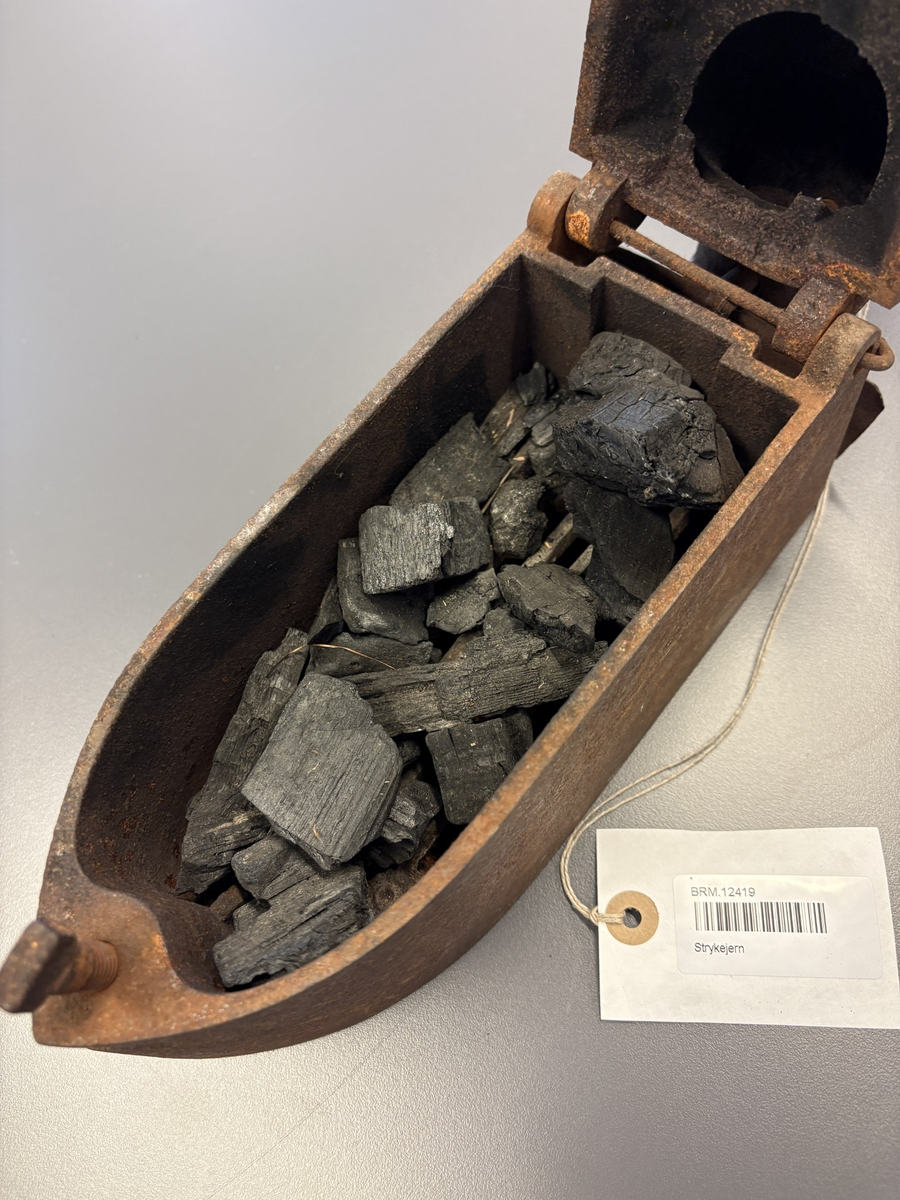 Båtformet strykejern av støpejern. Hengslet lokk med trehåndtak og pipe.