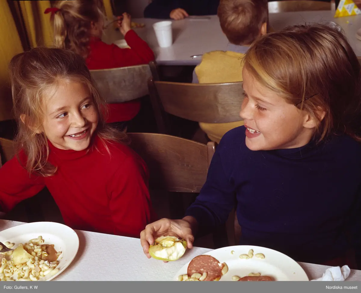 Två flickor äter lunch i skolbespisningen. Det serveras makaroner, korv och äpple.