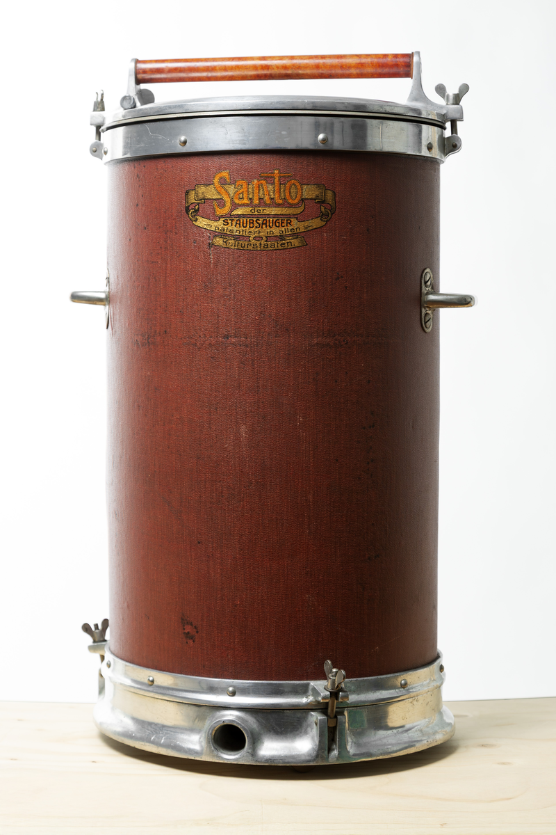 Stående cylindrisk dammsugare. Topplock av metall och cylinder, klädd med röd-brun pegamoid. Dammsugaren väger ca. 20 kg och är utrustad med både motor och pump.