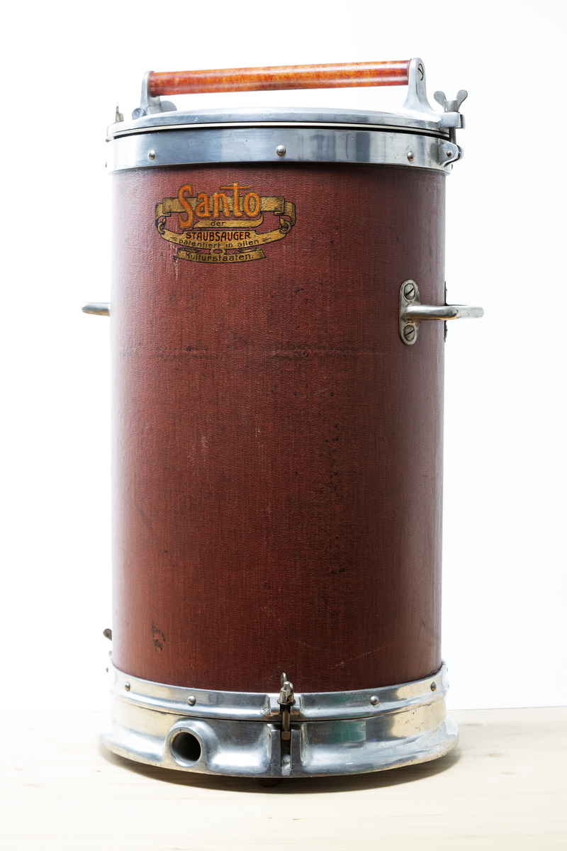 Stående cylindrisk dammsugare. Topplock av metall och cylinder, klädd med röd-brun pegamoid. Dammsugaren väger ca. 20 kg och är utrustad med både motor och pump.