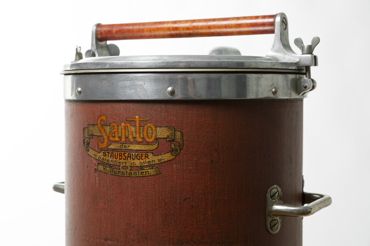 Stående cylindrisk dammsugare. Topplock av metall och cylinder, klädd med röd-brun pegamoid. Dammsugaren väger ca. 20 kg och är utrustad med både motor och pump.