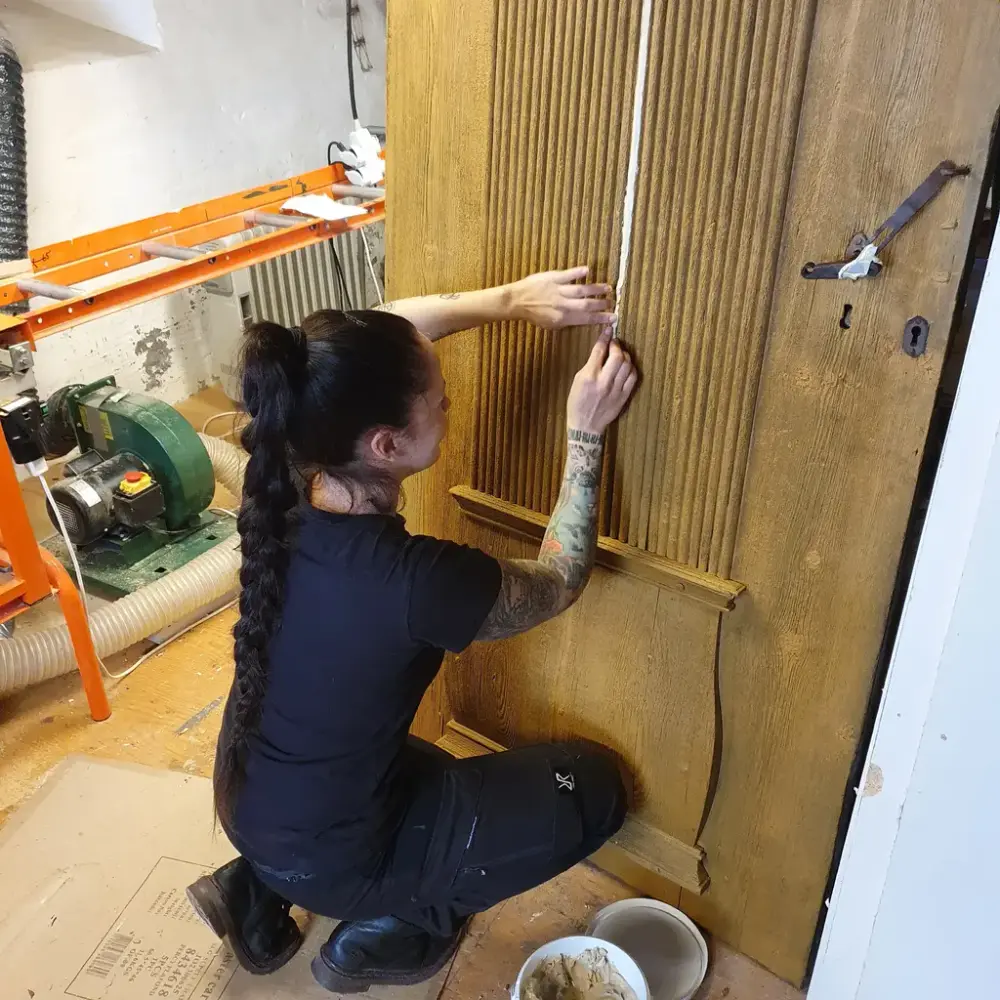 Kvinnelig museumsåndverker jobber med kitting av en gammel dør på verkstedet. 