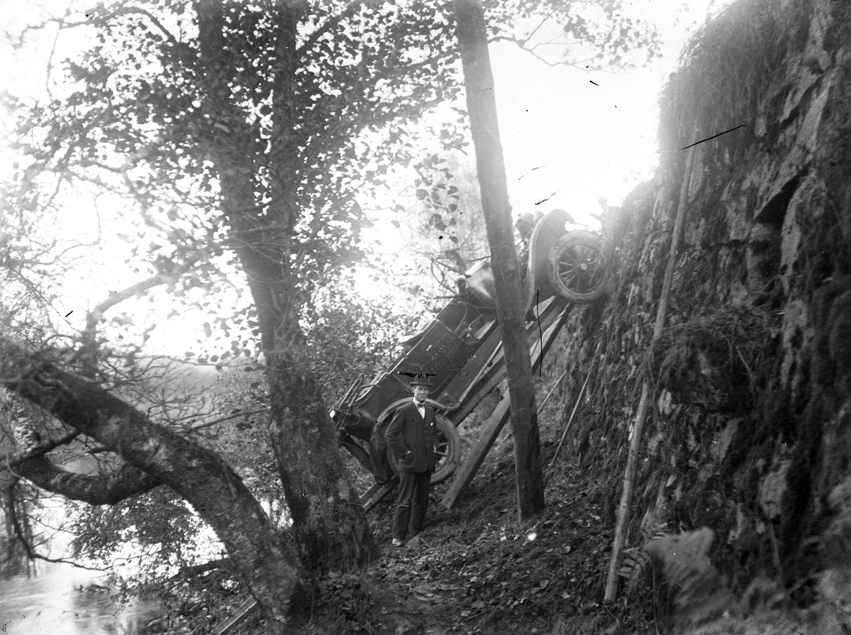 En bil har backat ner för en bergskant vid "Kleva", Säveån. Framför står en man och ler mot kameran. 
