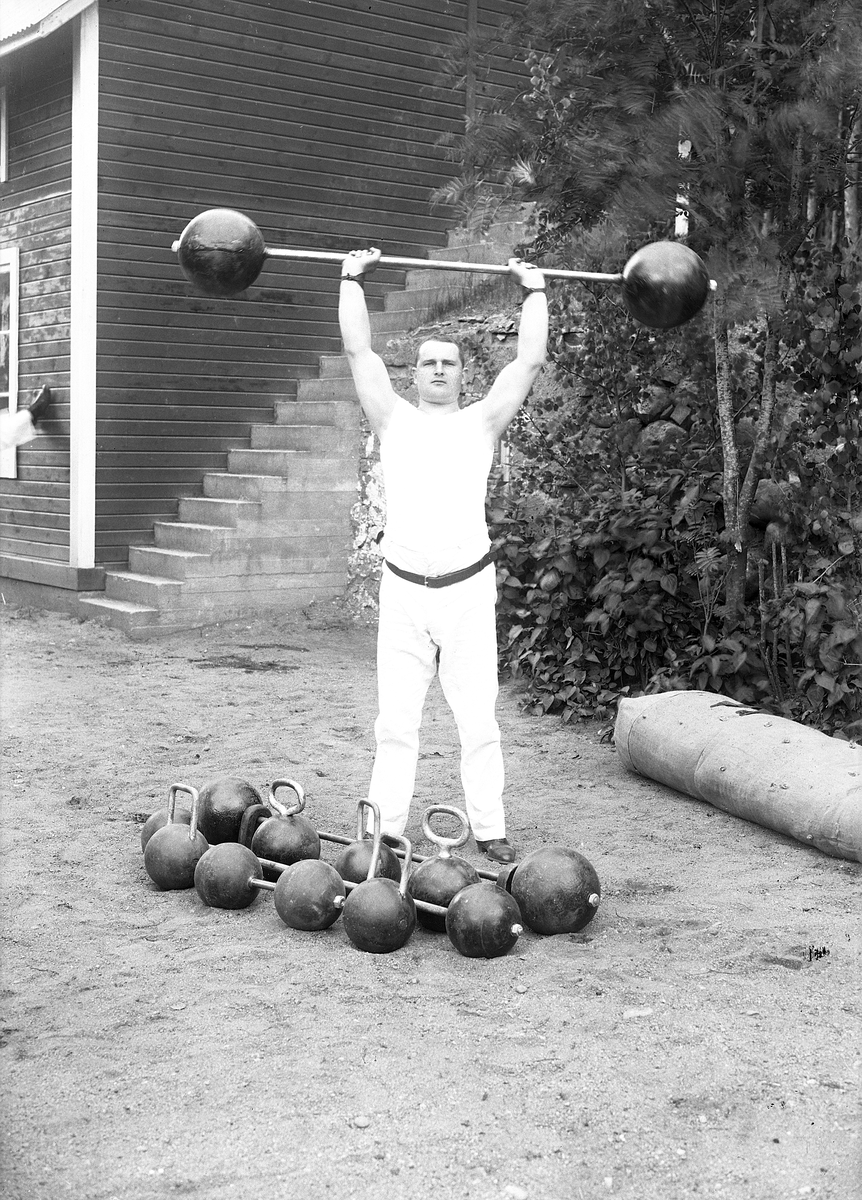 Kraftkarl ifrån atletklubben Heros poserar med viktstång av äldre modell försedd med klotformade järnvikter. Utomhus under tidigt 1900-tal.