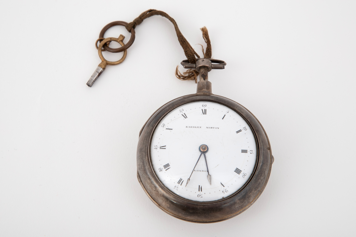 Lommeur med nøkkel. Lommeur med dobbel, glatt kasse av sølv. Innerkasse med gravert og gjenneombrutt dekor; mønsterbord og bladranker. Urskive i hvit emalje, med romerske timetall og arabiske minuttall. To stålvisere. Urnøkkel festet med en lisse til uret.