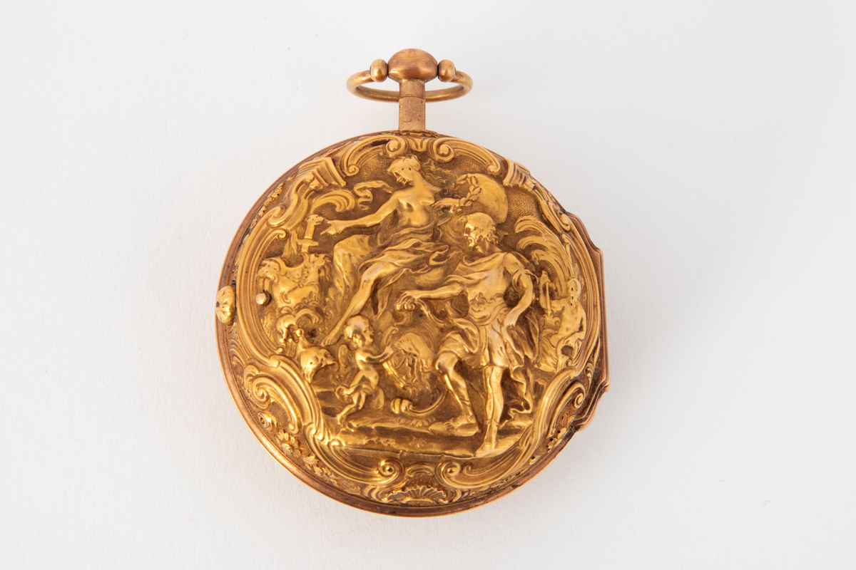 Spindelur med dobbel kasse i forgylt kobber. Ytterkasse med høyt relieff med figurscene: kvinne og mann med putto. Omramming med kartusjer og blomsterranker. Lignende kantbord på forsiden. Hvit emaljeskive med romerske timetall, arabiske minuttall og stålvisere.