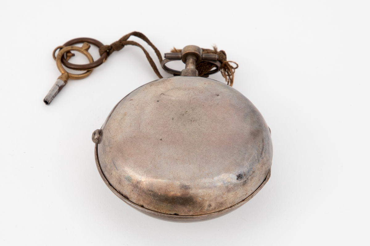 Lommeur med nøkkel. Lommeur med dobbel, glatt kasse av sølv. Innerkasse med gravert og gjenneombrutt dekor; mønsterbord og bladranker. Urskive i hvit emalje, med romerske timetall og arabiske minuttall. To stålvisere. Urnøkkel festet med en lisse til uret.