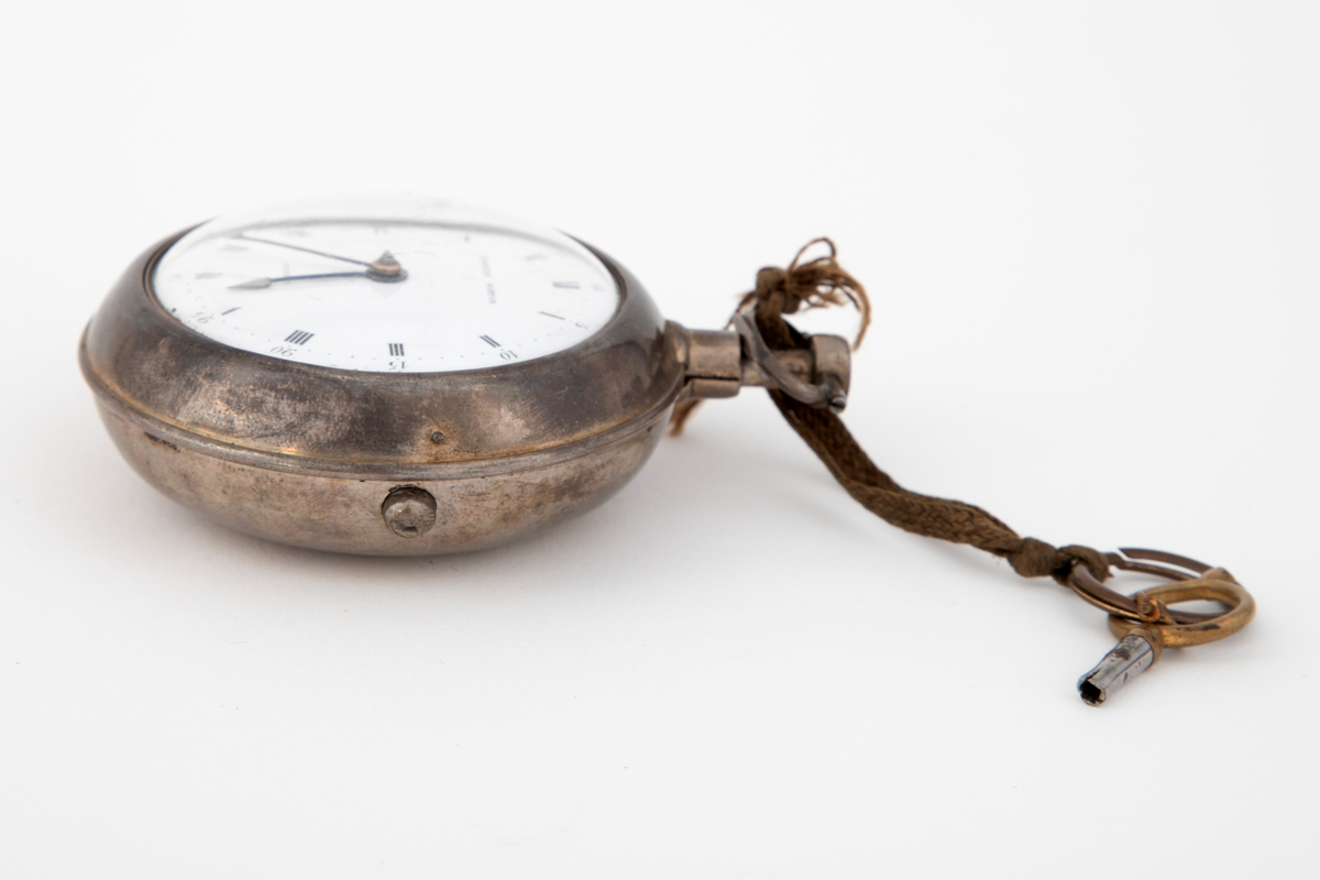 Lommeur med nøkkel. Lommeur med dobbel, glatt kasse av sølv. Innerkasse med gravert og gjenneombrutt dekor; mønsterbord og bladranker. Urskive i hvit emalje, med romerske timetall og arabiske minuttall. To stålvisere. Urnøkkel festet med en lisse til uret.