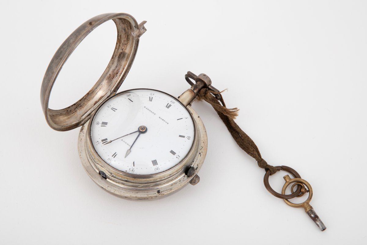Lommeur med nøkkel. Lommeur med dobbel, glatt kasse av sølv. Innerkasse med gravert og gjenneombrutt dekor; mønsterbord og bladranker. Urskive i hvit emalje, med romerske timetall og arabiske minuttall. To stålvisere. Urnøkkel festet med en lisse til uret.