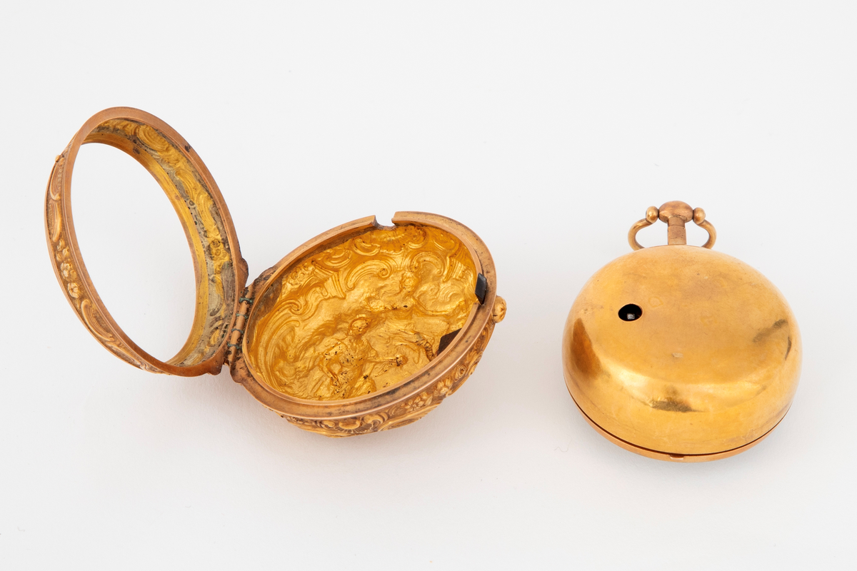Spindelur med dobbel kasse i forgylt kobber. Ytterkasse med høyt relieff med figurscene: kvinne og mann med putto. Omramming med kartusjer og blomsterranker. Lignende kantbord på forsiden. Hvit emaljeskive med romerske timetall, arabiske minuttall og stålvisere.