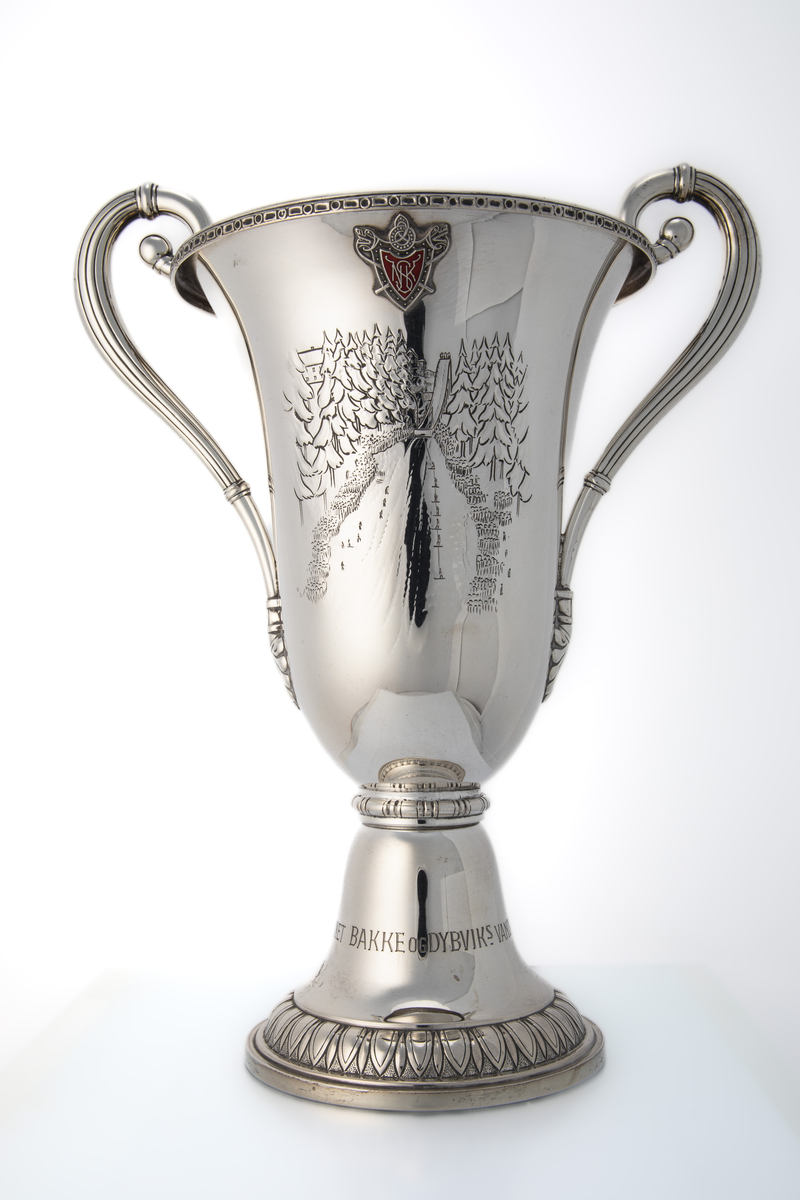Pokal med hanker og kort stett. På ene siden er det innskrift med navnene til hopperne på vinnerlagene. På andre siden er den dekorert med motiv som forestiller hoppbakken (Nydalsbakken) med publikum på begge sider, hoppere på vei opp med heis og en hopper i svevet. Over dette motivet er det et emblem (Nydalen Skiklubb).