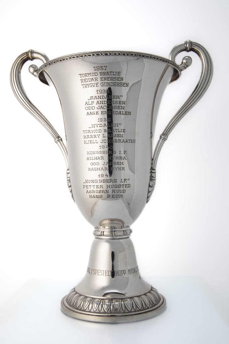 Pokal med hanker og kort stett. På ene siden er det innskrift med navnene til hopperne på vinnerlagene. På andre siden er den dekorert med motiv som forestiller hoppbakken (Nydalsbakken) med publikum på begge sider, hoppere på vei opp med heis og en hopper i svevet. Over dette motivet er det et emblem (Nydalen Skiklubb).