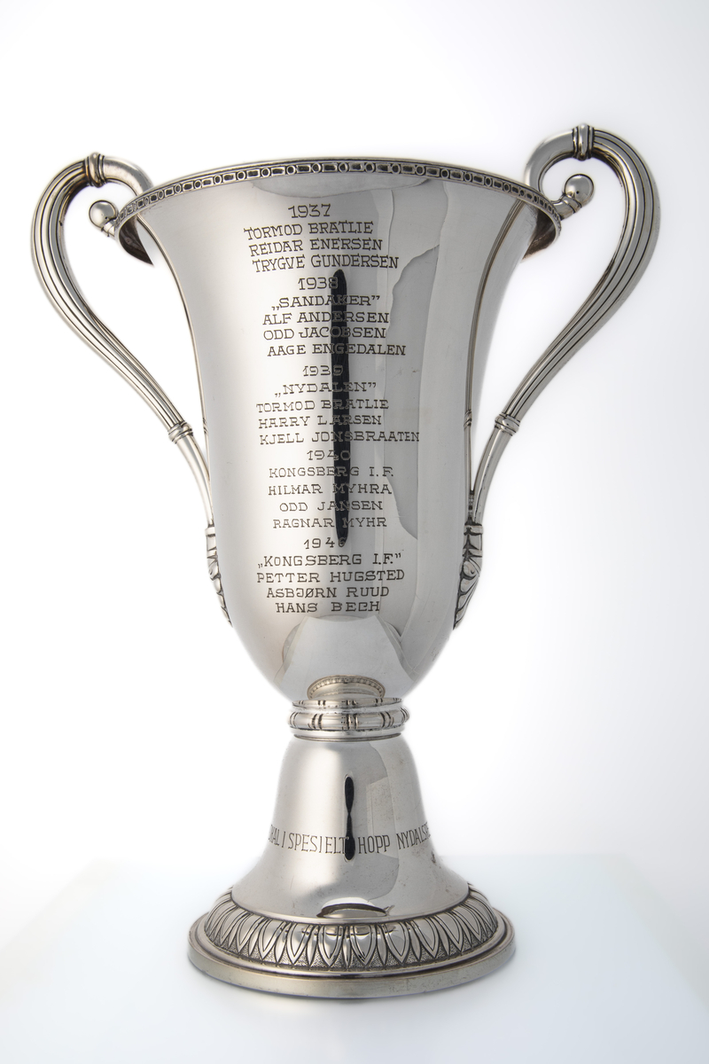 Pokal med hanker og kort stett. På ene siden er det innskrift med navnene til hopperne på vinnerlagene. På andre siden er den dekorert med motiv som forestiller hoppbakken (Nydalsbakken) med publikum på begge sider, hoppere på vei opp med heis og en hopper i svevet. Over dette motivet er det et emblem (Nydalen Skiklubb).
