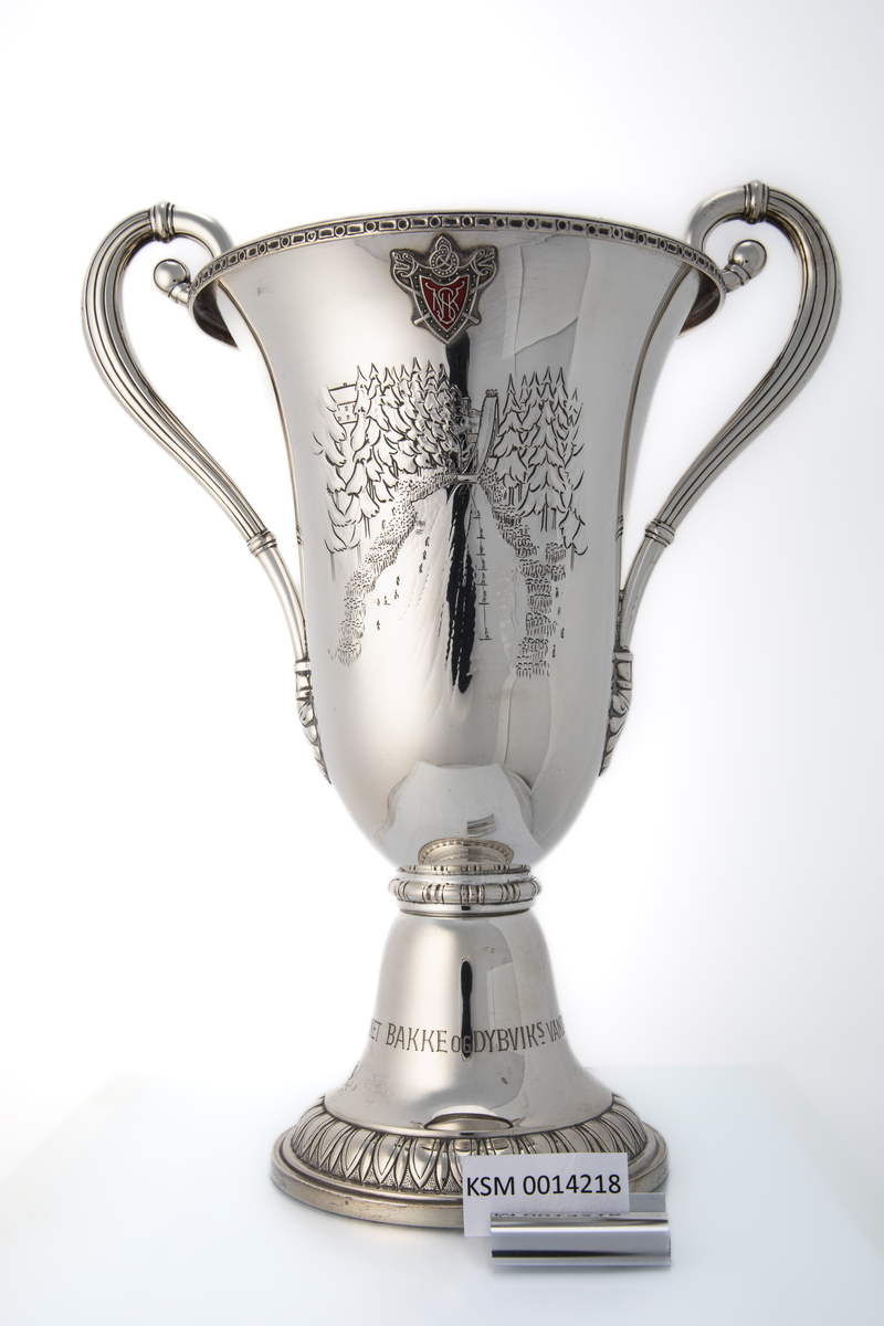 Pokal med hanker og kort stett. På ene siden er det innskrift med navnene til hopperne på vinnerlagene. På andre siden er den dekorert med motiv som forestiller hoppbakken (Nydalsbakken) med publikum på begge sider, hoppere på vei opp med heis og en hopper i svevet. Over dette motivet er det et emblem (Nydalen Skiklubb).