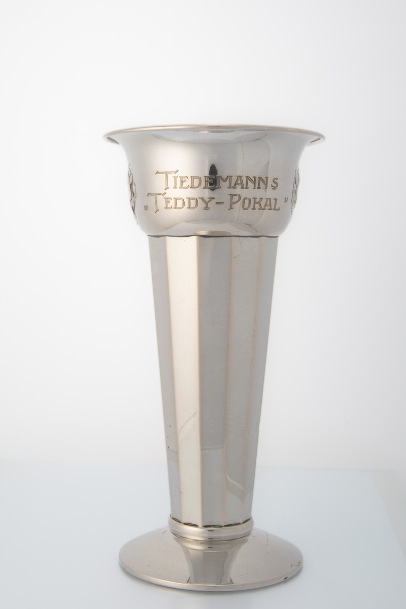 Sølvpokal med mangekanta form på hoveddelen, låg stett og motiv og innskrift på øvre del. Motiv av hovudet til ein tiger (Tiedemanns tobakk logo).