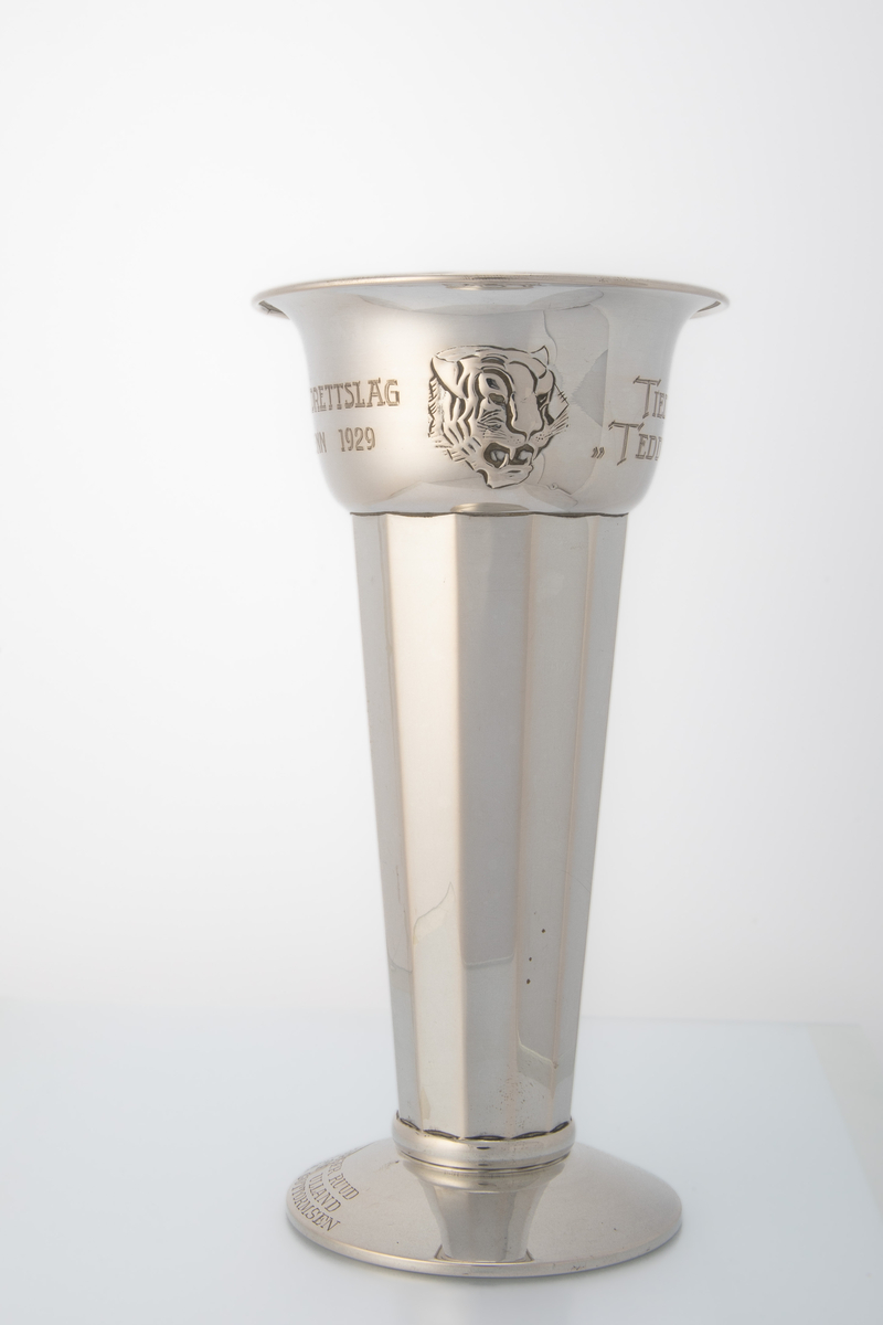 Sølvpokal med mangekanta form på hoveddelen, låg stett og motiv og innskrift på øvre del. Motiv av hovudet til ein tiger (Tiedemanns tobakk logo).