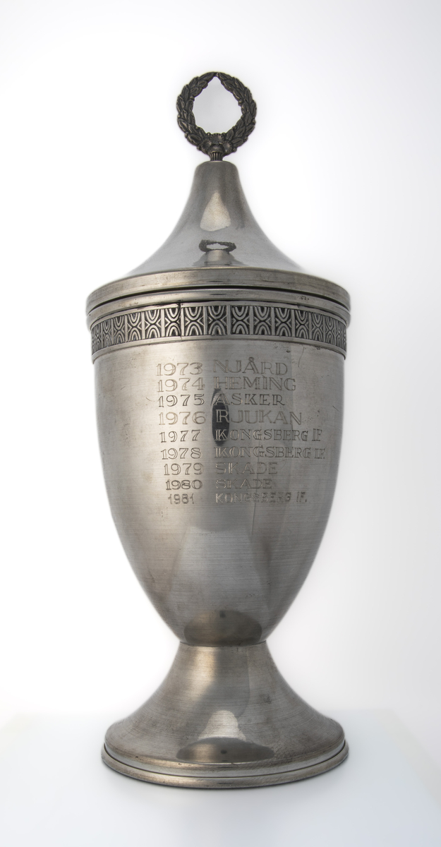 Tinnpokal med lokk og stett. På begeret er det emblem (Rjukan Idrettslag), inskripsjon og en dekorativ mønsterbord langs kanten øverst. Lokket har en spiss form. Vandrepokal for Rjukanrennet, gutter og jenter, lag.