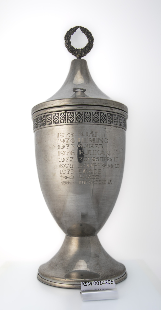 Tinnpokal med lokk og stett. På begeret er det emblem (Rjukan Idrettslag), inskripsjon og en dekorativ mønsterbord langs kanten øverst. Lokket har en spiss form. Vandrepokal for Rjukanrennet, gutter og jenter, lag.