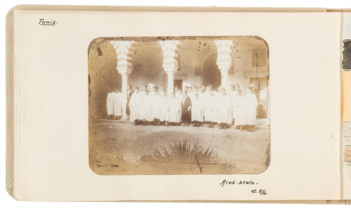 Denna fotografi visar en grupp unga män i arabiska kläder som står framför en byggnad i morisk stil.