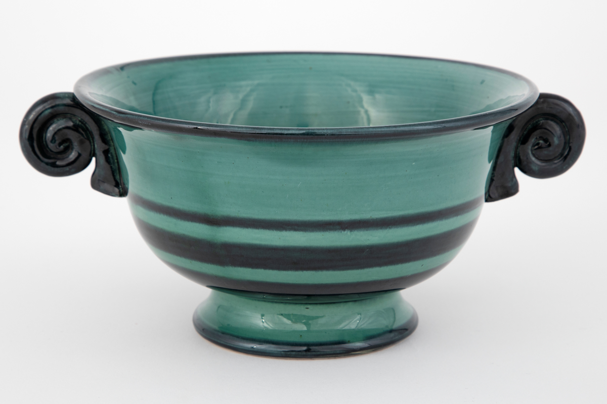 Vase i fajanse. Konkav fot, rund korpus, svakt utoverbrettet kant samt spiralformede hanker. Vasen er glasert med en blank, grønn glasur. Dekorert med horisontale, svarte ringer i forskjellige tykkelser. Hankene er glasert med en blank, svart glasur. Tett, finkornet hvitt gods.