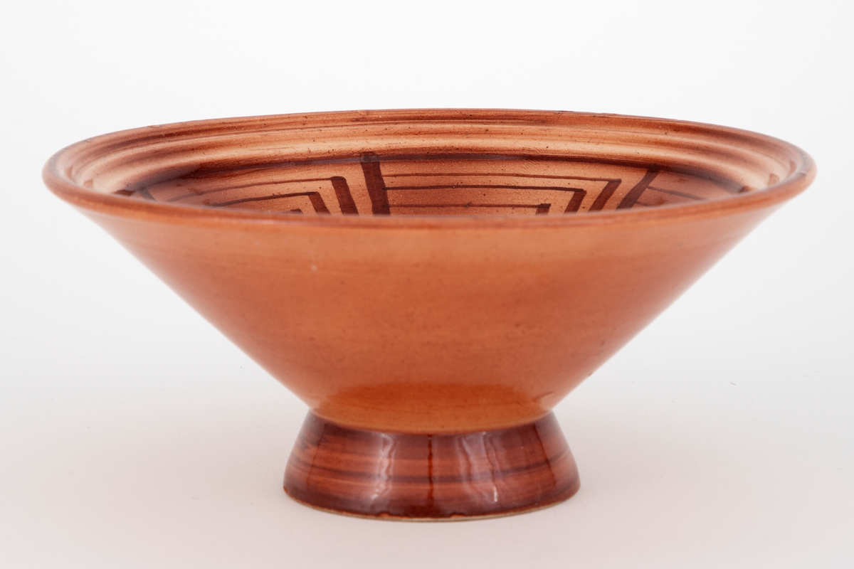Skål i fajanse. Konkav fot og kjegleformet korpus. Skålen er glasert med en blank, rød glasur. Innsiden er dekorert med et geometrisk mønster i mørkerødt. Tett, finkornet hvitt gods.