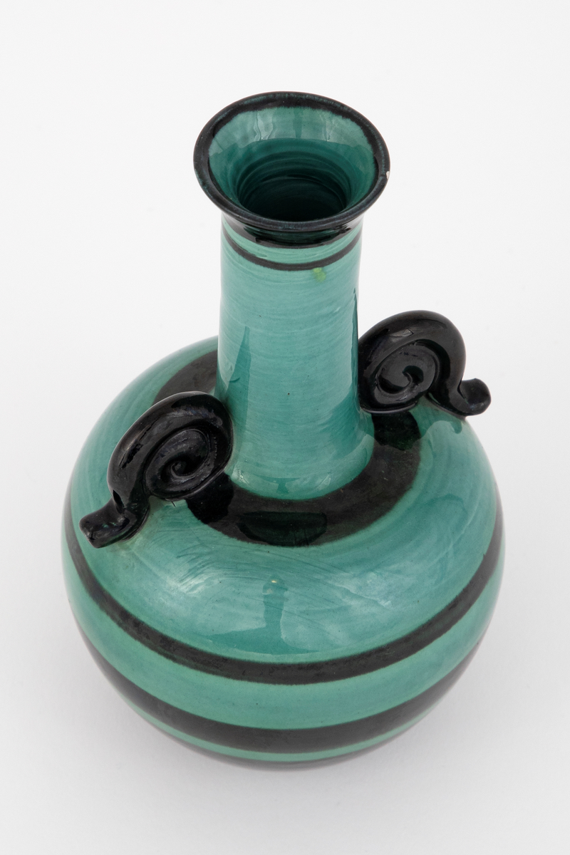 Vase i fajanse. Kuleformet korpus med en lang, smal hals og en svak, utoverbrettet munning. Spiralformede hanker der korpus går over til hals. Vasen er glasert med en blank, grønn glasur. Dekorert med horisontale, svarte striper i forskjellige tykkelser. Tett, finkornet hvitt gods.