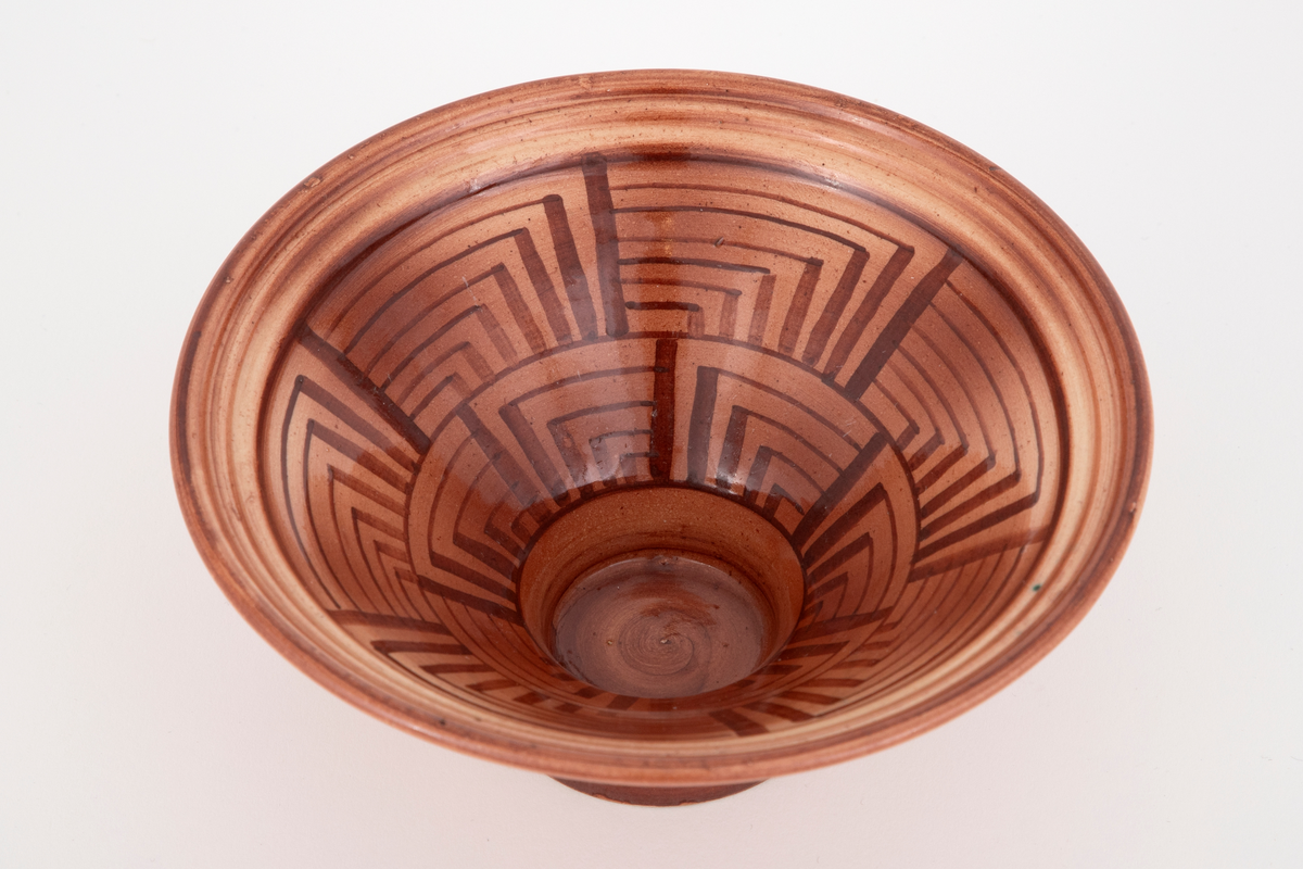 Skål i fajanse. Konkav fot og kjegleformet korpus. Skålen er glasert med en blank, rød glasur. Innsiden er dekorert med et geometrisk mønster i mørkerødt. Tett, finkornet hvitt gods.