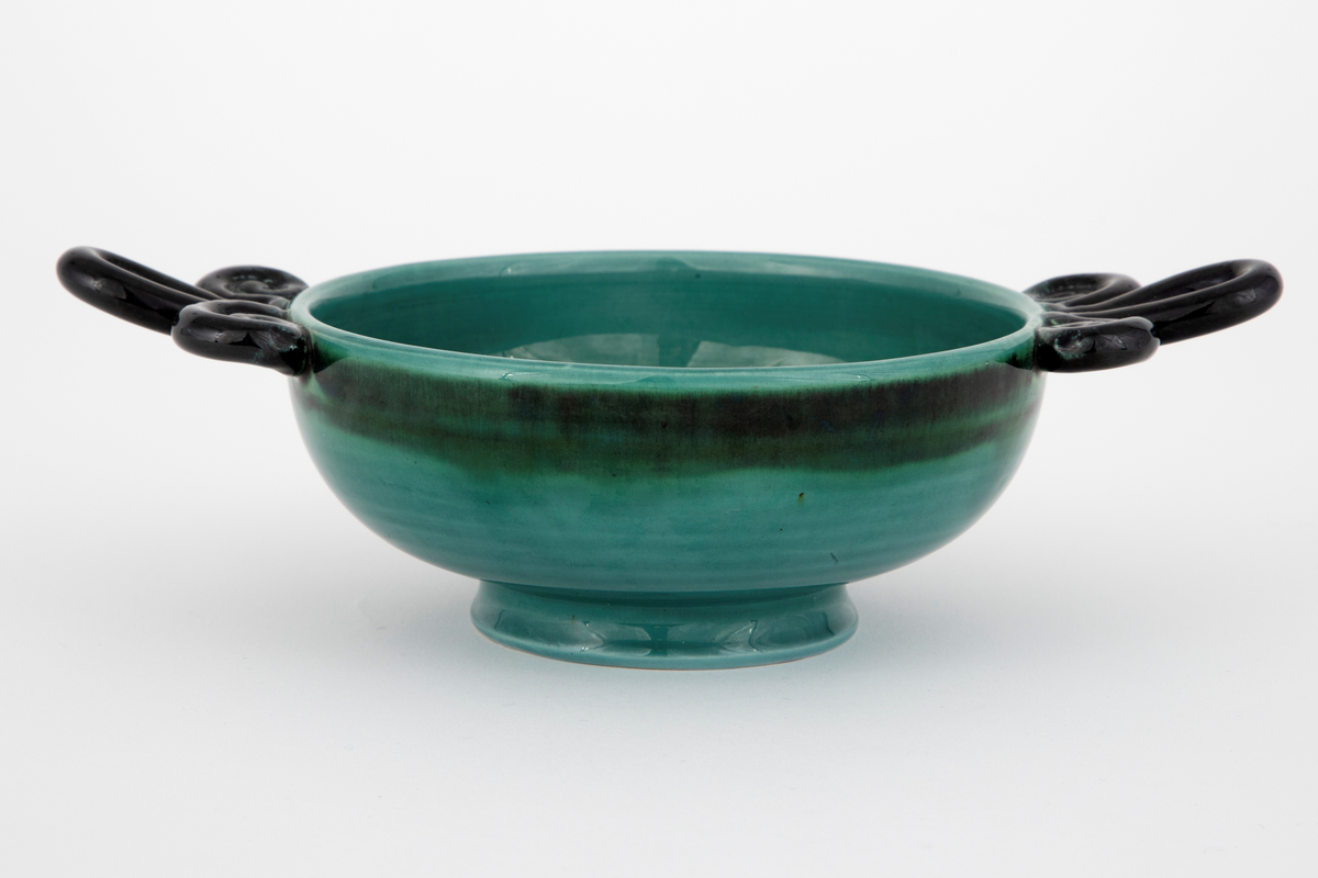 Skål i fajanse. Lav, konkav fot, halvkuleformet korpus og spiralformede hanker. Skålen er glasert med en blank, grønn glasur som er mørkere langs kanten. Innsiden er dekorert med horisontale, svarte ringer i forskjellige tykkelser. Hankene er glasert med en blank, svart glasur. Tett, finkornet hvitt gods.