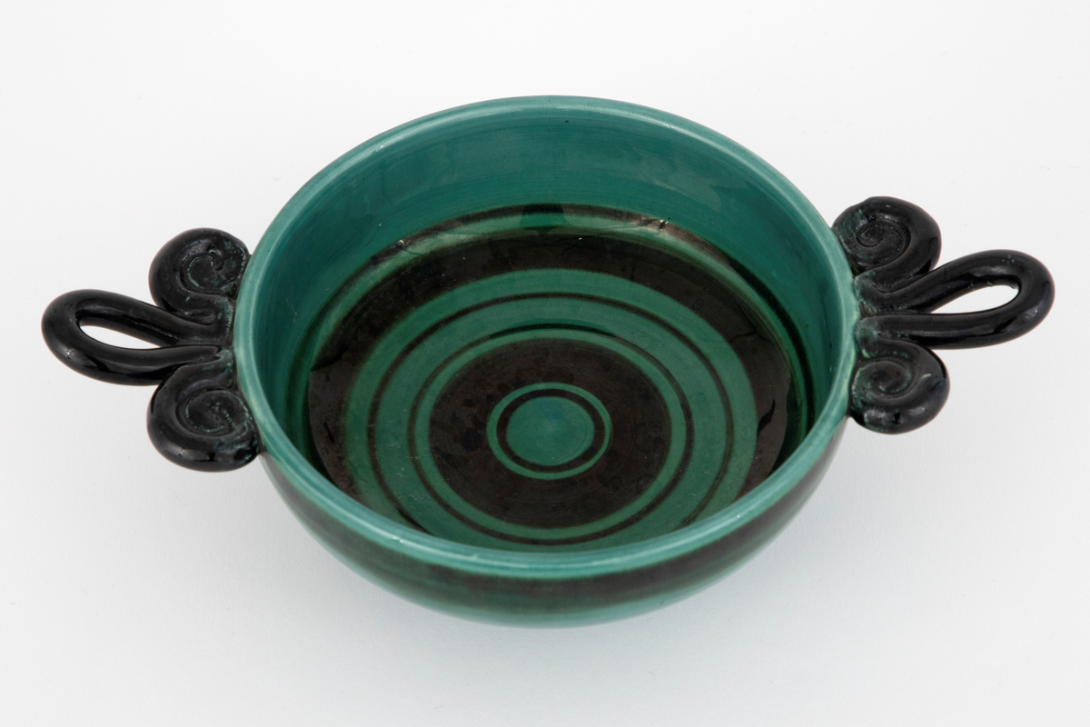 Skål i fajanse. Lav, konkav fot, halvkuleformet korpus og spiralformede hanker. Skålen er glasert med en blank, grønn glasur som er mørkere langs kanten. Innsiden er dekorert med horisontale, svarte ringer i forskjellige tykkelser. Hankene er glasert med en blank, svart glasur. Tett, finkornet hvitt gods.