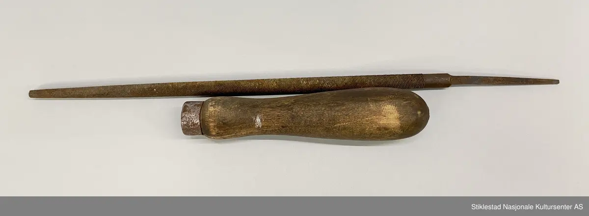 Fabrikkprodusert fil med dreid skaft av løvtre. Fila er rund og smalner av mot enden, dobbelthogd. Holk i jernblikk. Innsnevring i nedre del av skaft for bedre grep. En god del rust, skaftet er sprukket, fil løs fra skaft. Produksjonsmerke, men ikke lesbart.