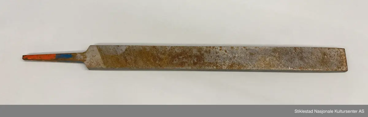 Fabrikkprodusert fil uten skaft. Dobbelhogd på begge sider (skråstilt rutenett). Kortsider har rette horisontale hogg. Rester av rød og blå farge på tange. Stemplet JM 13? på ene siden. Andre siden: Vilala-Finland. Rust.