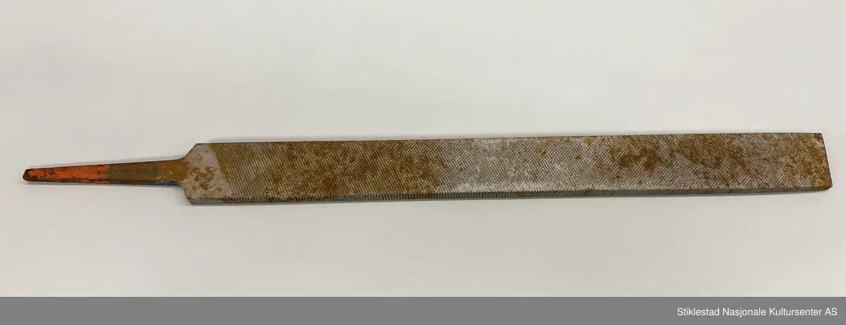 Fabrikkprodusert fil uten skaft. Dobbelhogd på begge sider (skråstilt rutenett). Kortsider har rette horisontale hogg. Rester av rød og blå farge på tange. Stemplet JM 13? på ene siden. Andre siden: Vilala-Finland. Rust.