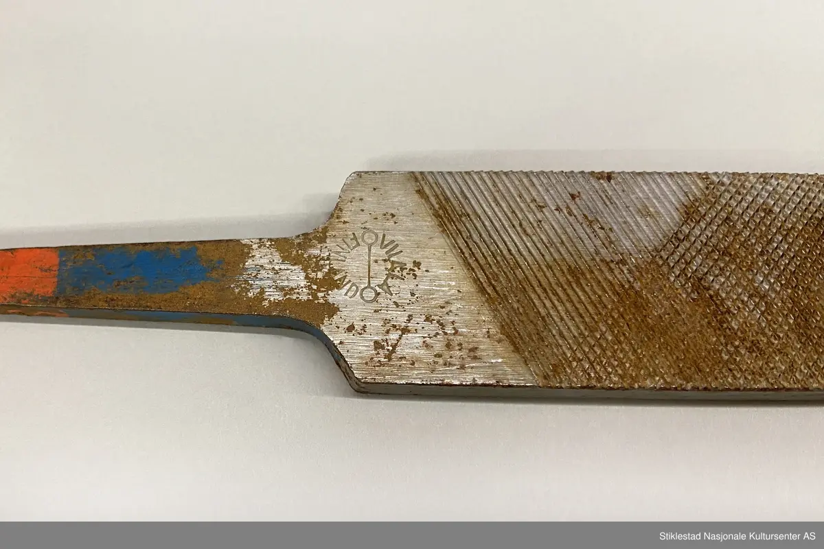 Fabrikkprodusert fil uten skaft. Dobbelhogd på begge sider (skråstilt rutenett). Kortsider har rette horisontale hogg. Rester av rød og blå farge på tange. Stemplet JM 13? på ene siden. Andre siden: Vilala-Finland. Rust.