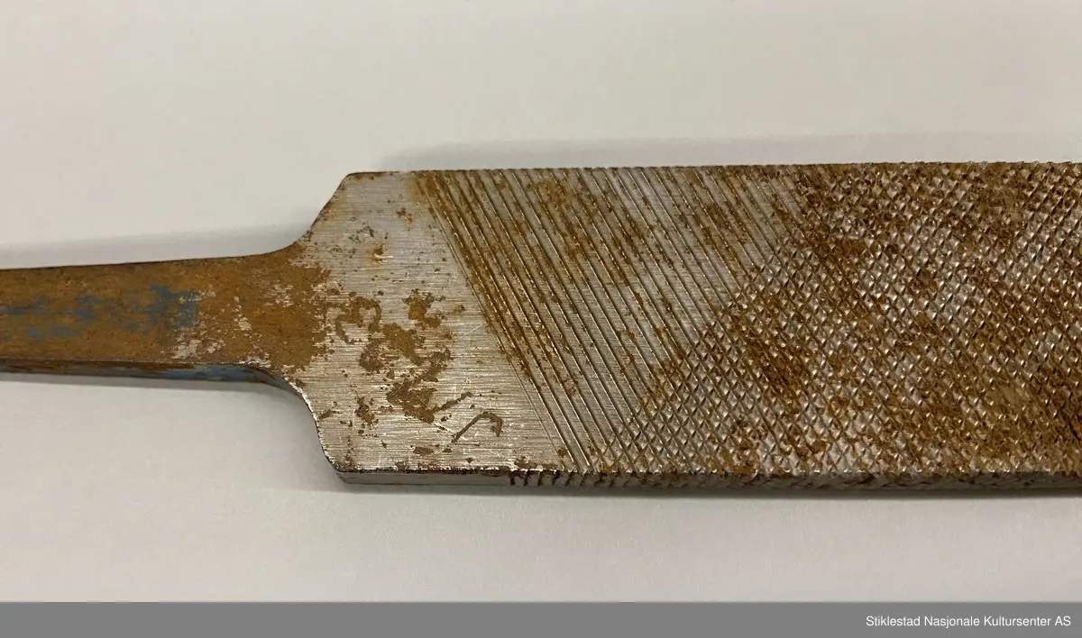 Fabrikkprodusert fil uten skaft. Dobbelhogd på begge sider (skråstilt rutenett). Kortsider har rette horisontale hogg. Rester av rød og blå farge på tange. Stemplet JM 13? på ene siden. Andre siden: Vilala-Finland. Rust.