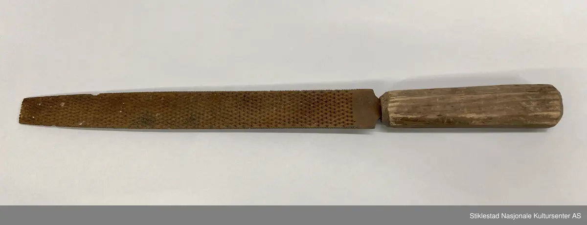 Fabrikkprodusert rasp med teljet/spikket skaft av løvtre. Raspa er flat på en side og buet på motsatt side, smalner av mot enden, spissen er knekt. På buet side av fila, ser enden ut til å være (delvis) slipt bort. En god del rust.