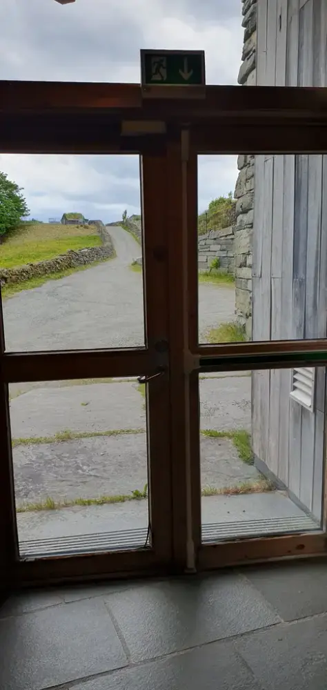 Ytterdør og naudutgang i glass, frå auditoriet. Grusbakken opp til hovudvegen i bakgrunnen.