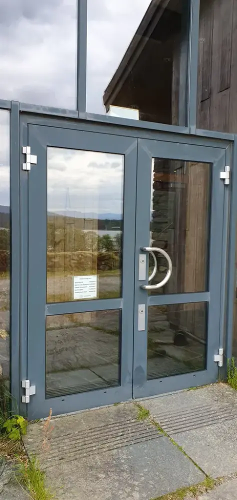 Dobbel ytterdør i glass, med vidare tilkomst til lager og gangen i underetasjen. Døra er på nedsida av informasjonsbygget.