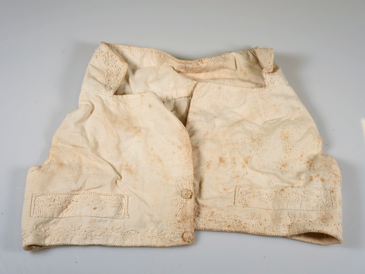 Handsydd barnväst i linne och bomull. Mycket smalt ryggstycke. Dekorerad med broderi i vitt längs krage, fickor och västens nederkant. Även de två klädda knapparna är dekorerade med broderi.