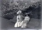 Två kvinnor (en äldre och en yngre) med stora hattar, har slagit sig ner på en parksoffa, troligen vid strandpromenaden i Varberg.