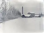 En vy över en fabrik (tegelbruk?), med en åker i förgrunden. 
Vinterbild.