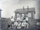 Några sommarklädda barn och vuxna har samlats kring ett enkelt kaffebord utomhus. I bakgrunden syns ett bostadshus, troligtvis hyrt som sommarställe.