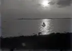 Landskap med hav och en segelbåt i motljus. I bakgrunden syns en fyr.