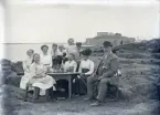 En familj (?) har samlats i det fria, kring ett bord med saft och vatten m.m. I bakgrunden syns Varbergs fästning.