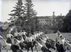 En trupp kvinnliga gymnaster övar i det fria utanför dåv. seminariet, dvs numera Ringsbergsskolan i Växjö.  I bakgrunden syns skolans växthus och dåv. cellfängelset.